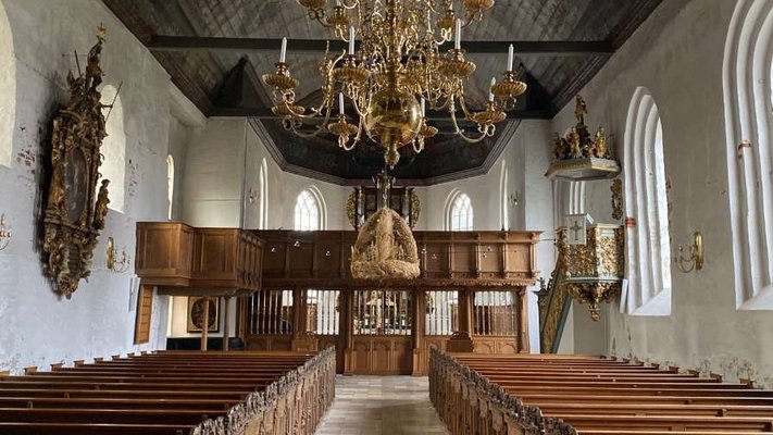 Es ist die St. Laurentius Kirche von innen zu sehen. Das Motiv zeigt einen Gang zwischen zwei Bankreihen. Von der verzierten Decke hängt ein Kronleuchter.