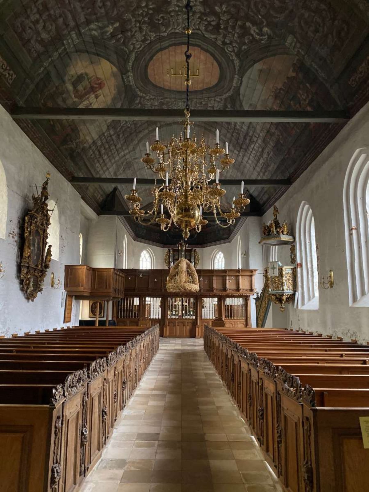 Es ist die St. Laurentius Kirche von innen zu sehen. Das Motiv zeigt einen Gang zwischen zwei Bankreihen. Von der verzierten Decke hängt ein Kronleuchter.